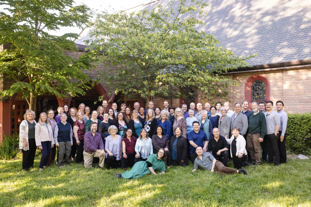 members of the Seattle Choral Company in business casual clothing, on the lawn in front of Phinney Ridge Lutheran church. Two trees and the brick exterior of the church are in the background.